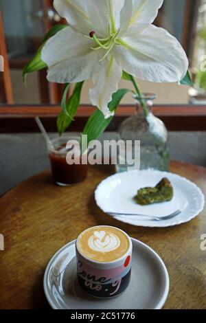 Tazza di ceramica di latte caldo, torta di tè verde Matcha, un bicchiere di americano ghiacciato su tavolo di legno decorato con fiore di giglio Foto Stock