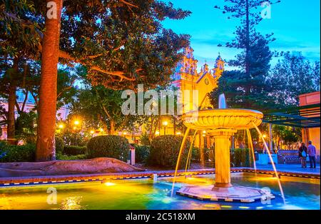 La serata in Alameda Apodaca e nel giardino Marques de Comillas con vista sulla fontana di pietra d'epoca e la chiesa Carmen illuminata sullo sfondo, Cadice Foto Stock