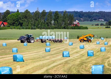 Ambiente rurale con campo di fieno e raggiungere il carrello elevatore impilando balle di insilato sul trattore trainato rimorchio agricolo in una bella giornata d'estate. Foto Stock