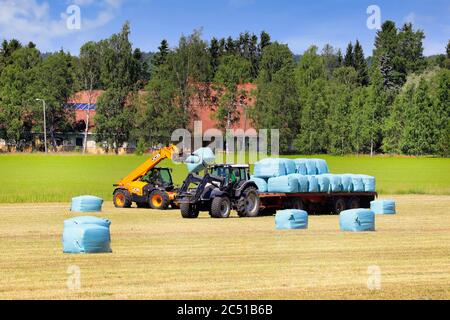 Ambiente rurale con campo di fieno e carrello elevatore JCB che impilano balle di insilato su un rimorchio agricolo trainato da un trattore Valtra. Salo, Finlandia. 27 giugno 2020. Foto Stock