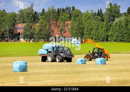 Ambiente rurale con campo di fieno e carrello elevatore JCB che impilano balle di insilato su un rimorchio agricolo trainato da un trattore Valtra. Salo, Finlandia. 27 giugno 2020. Foto Stock