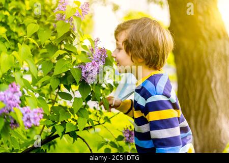 Ragazzo felice che togli la maschera medica protettiva e respirando aria, annusare fiori di lilla. Allegro bambino caucasico al sole giorno all'aperto nel parco. Foto Stock