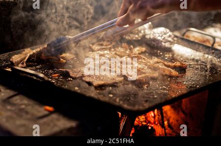 Fumo e vapore che si alzano da sottili strisce di carne che vengono grigliate su una grande pentola di metallo su un fuoco di legno Foto Stock