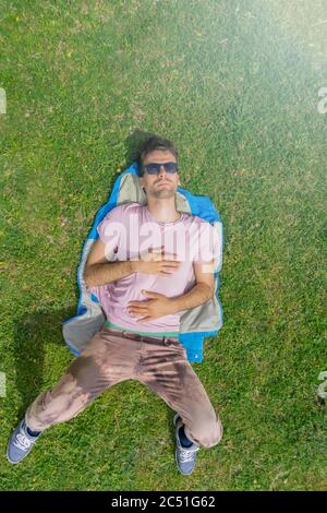 Uomo attraente adagiato su erba verde, rilassante calda giornata estiva. Vista dall'alto del ragazzo che riposa sull'erba. Disposizione piatta con spazio per la copia. Concetto di relax estivo. Vertic Foto Stock