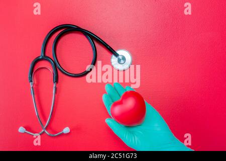 Stetoscopio medico e mano in guanto di gomma verde che tiene il cuore rosso su sfondo rosso. Posa piatta di cuore isolato in mano in guanto e stetoscopio Foto Stock