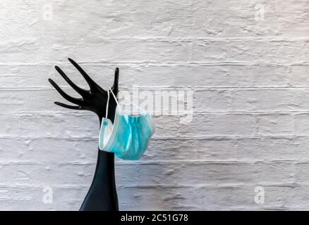 Mano di plastica nera che tiene la maschera medica monouso sul fondo bianco della parete di mattoni. Manichino in plastica per gioielli per la protezione del viso facem Foto Stock