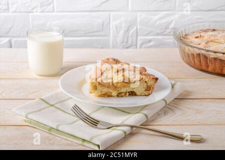 Un pezzo di torta di mele dolce e acido su un piatto bianco e un bicchiere di latte su un tavolo di legno bianco. Ricetta per dolci e dolci fatti in casa. Pasticceria per la colazione Foto Stock
