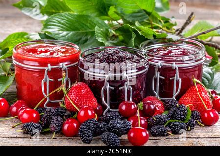 Fragola, ciliegia e marmellata di gelsi su un tavolo rustico, con una varietà di frutti di bosco freschi come decorazione. Foto Stock