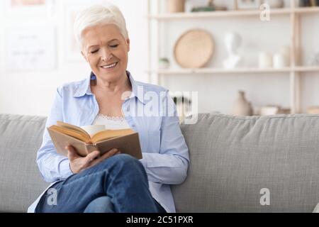 Passatempo preferito. Sorridente anziano Signora leggere Libro su comodo divano a casa Foto Stock