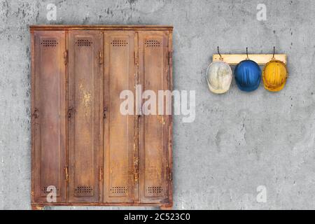 Armadietto in arrugginito d'epoca e caschi da costruzione appesi su un portabicchiette di fronte a una vecchia parete grigia Foto Stock