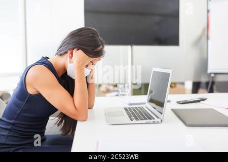 Donna di affari che indossa la maschera facciale a causa di Covid-19 si siede con burnout dovuto sovraccarico sul computer Foto Stock