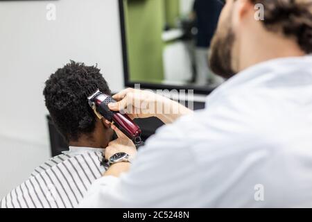 Vista laterale dell'uomo con elegante e moderno taglio di capelli che guarda in avanti nel barbiere. Mano di barbiere che tiene il rasoio e tagliando i capelli sulla testa del cliente. Concetto Foto Stock