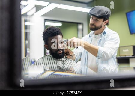 Felice giovane uomo nero che viene rifinito con la macchina elettrica del regolacapelli in barbiere. Concetto di trattamento di bellezza maschile. Sorridente giovane africano che si sta facendo nuovo Foto Stock