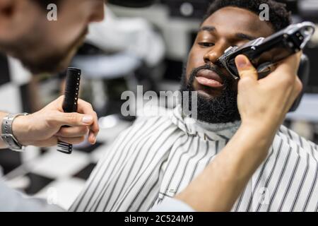 Barbiere rifinire i capelli con il regolacapelli su giovane uomo nero unshaven in barbershop studio.Professional parrucchiere tagliare i capelli con tagliatrice elettrica su Afri Foto Stock