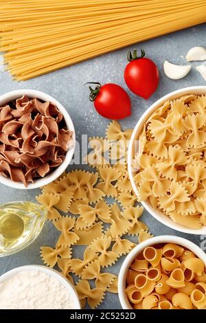 Varietà di pasta cruda di diverse forme e forme con pomodori, olio, aglio e farina. Vista dall'alto. Foto Stock