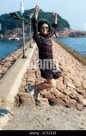 Una donna cinese che salta su un molo di pietra al largo dell'isola faro di xiao qingdao in Cina, provincia di Shandong. Foto Stock