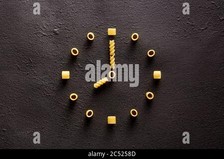 Pasta cruda disegnata a forma di orologio su sfondo marrone scuro. Mangiare la pasta nel concetto di cena. Foto Stock