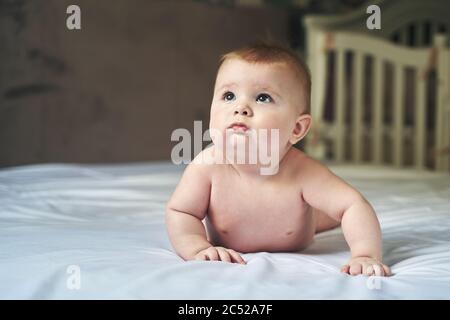 un bambino meraviglioso di sei mesi si trova su un grande letto su un foglio bianco e guarda in alto Foto Stock