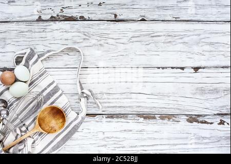 Grembiule con fornimenti di cottura. Frusta, misurini, cucchiaio di legno vecchio e uova su uno sfondo di legno bianco. Immagine ripresa dalla vista superiore. Foto Stock