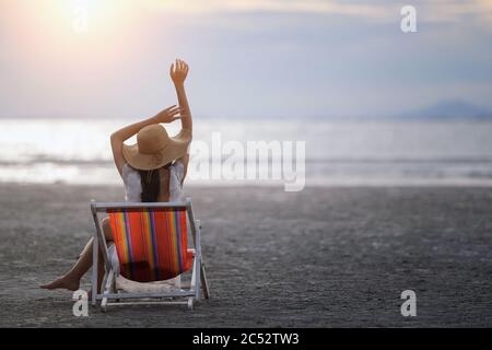 Vista posteriore di una donna seduta su una sedia a sdraio sulla spiaggia al tramonto, Thailandia Foto Stock