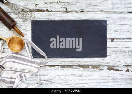 Cartello bianco con prodotti da forno. Grembiule, frusta, cucchiaio vecchio di legno e antica spilla su uno sfondo o un tavolo di legno bianco. Immagine ripresa dalla parte superiore del vie Foto Stock