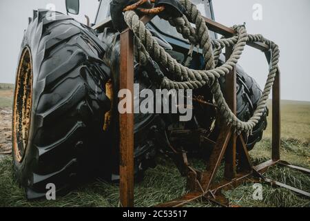 Dettaglio della spessa fune umida avvolta e appesa al telaio posteriore arrugginito sulla parte posteriore di un vecchio trattore abbandonato con enormi ruote doppie Foto Stock