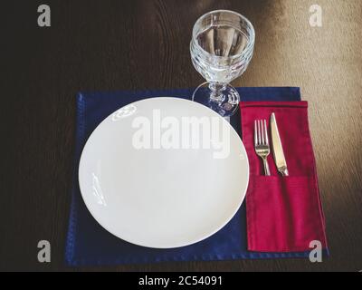 Una piastra in ceramica bianca con un vetro e argenteria giace su un tovagliolo su una superficie di legno. Impostazione del tavolo Foto Stock