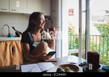 Giovane donna seduta su un tavolo da cucina in legno con atmosfera rilassata controllando il suo smartphone e scrivere note; concetto di stile di vita naturale, semplicità Foto Stock