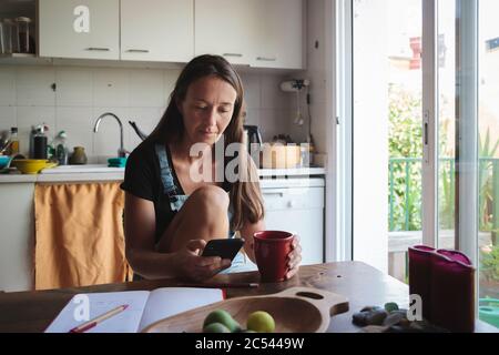 Giovane donna seduta su un tavolo da cucina in legno con atmosfera rilassata controllando il suo smartphone e scrivere note; concetto di stile di vita naturale, semplicità Foto Stock