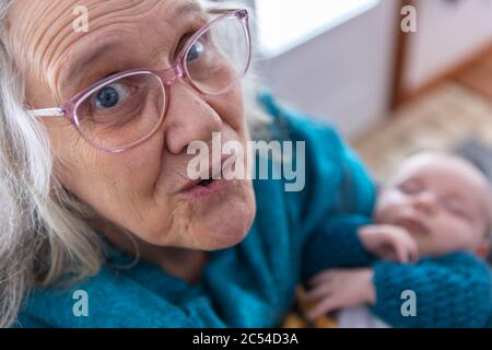 Ritratto a fuoco selettivo della donna anziana in occhiali rosa guardando verso la fotocamera, fuori fuoco vediamo un bambino piccolo ondato tra le braccia Foto Stock