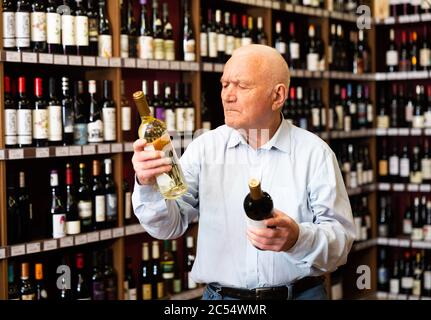 Un anziano serio sceglie tra vino rosso e bianco in un negozio di liquori Foto Stock