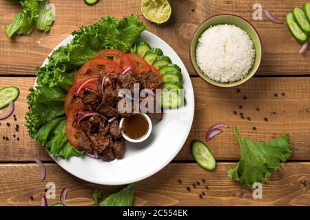 Piatto Bo Luc Lac su fondo di legno. Chiamato anche Lok Lak in cambogia o scuotendo manzo in America. Gustosi cubetti di manzo saltato marinato sono serviti con fr Foto Stock