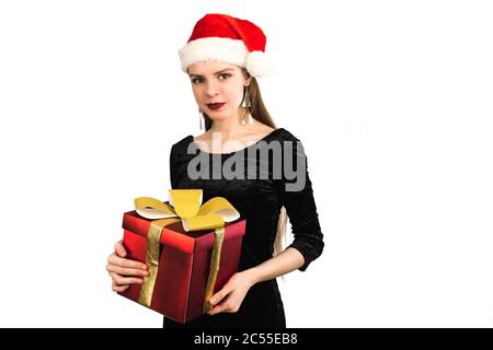 Bella ragazza che tiene dono e sorridendo alla macchina fotografica su sfondo bianco isolato. Un modello in un abito natalizio e cappello Babbo Natale danno un regalo rosso grande con un arco d'oro. Foto Stock