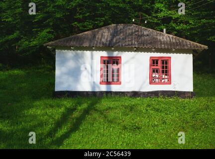 Casa bianca con finestre rosse. Antica casa di campagna al prato verde. Rifugio ai margini della foresta Foto Stock