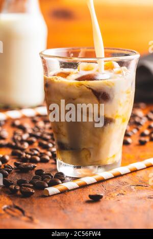 Versare il latte in un bicchiere con caffè ghiacciato su un tavolo di legno. Foto Stock