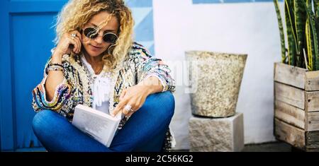 Bella donna adulto modello biondo godere il libro lettura all'aperto con porta blu e casa bianca in background - persone e attività di svago - alla moda Foto Stock