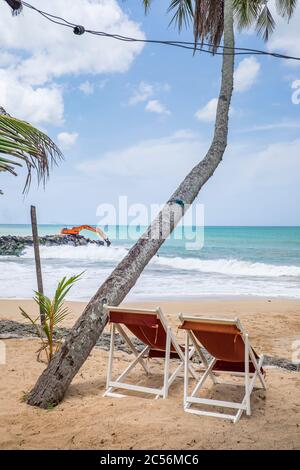 Sedie a sdraio sulla spiaggia di fronte al mare con gli escavatori su una scogliera sulla costa dello Sri Lanka Foto Stock