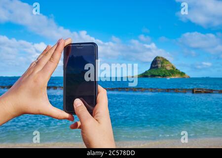 Donna scatta foto di Kualoa Rock con il suo smartphone sulla spiaggia, Kualoa Rock Beach, Kualoa Point, Isola di Mokoli'i, Parco Regionale di Kualoa, Hawaiiano Foto Stock