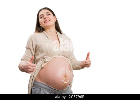 Madre futura sorridente e facendo un gesto di pollice-su con crema smiley faccia disegno sulla sua pancia isolato su sfondo bianco come sana gravidanza conce Foto Stock