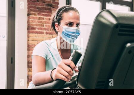 Giovane donna brunetta fa il ciclismo statico in palestra. Indossa una maschera. Foto Stock