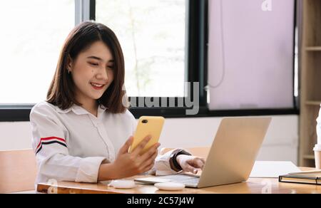 La ragazza asiatica lavora da casa o uno studente sta studiando da casa o da un freelance. Utilizza uno smartphone e un computer portatile Foto Stock