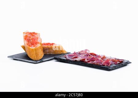 Deliziose fette di tipico prosciutto spagnolo su un piatto scuro accanto ad alcune fette di pane sfregate di pomodoro su un piatto scuro su sfondo chiaro. Foo tradizionale Foto Stock