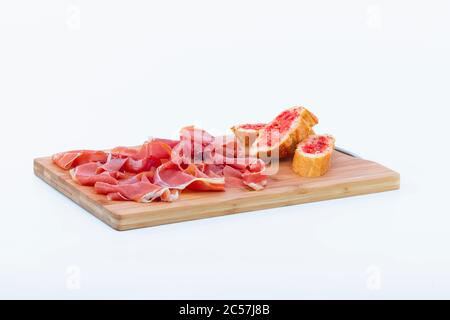 Deliziose fette di prosciutto spagnolo tipico accanto ad alcune fette di pane sfregate con pomodoro su un tagliere di legno su uno sfondo chiaro. Tradizionale f Foto Stock