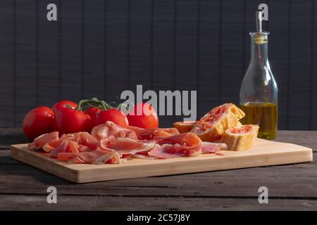 Sottili fette di prosciutto spagnolo tipico, pane con pomodoro, pomodori freschi e una bottiglia di vetro con olio d'oliva su un tavolo di legno su un backgro fuori fuoco Foto Stock