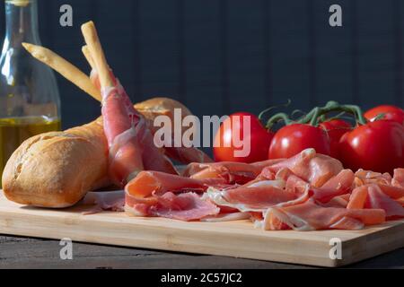 Primo piano di tipiche fette di prosciutto spagnolo, pomodori, bastoncini di pane avvolti in prosciutto e un pane su un tavolo di legno su uno sfondo fuori fuoco. Tradizionale Foto Stock