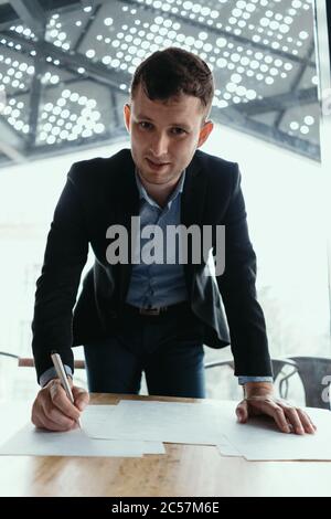 Giovane uomo d'affari sicuro che firma i documenti in un ufficio moderno con la finestra sullo sfondo. Penna in mano, carte sulla scrivania di legno Foto Stock