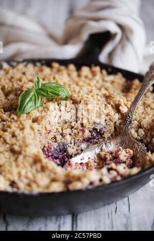 Dolce casa mora e mirtillo Basil Cobbler cotto in una padella di ghisa e sormontato con una farina d'avena dorata croccante su un rustico tavolo di legno bianco. Foto Stock