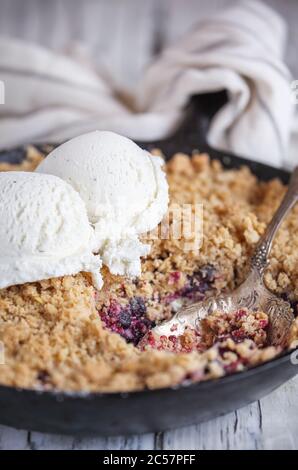 BlackBerry e Blueberry Cobbler cotti in una padella di ghisa e sormontati con una farinata d'avena dorata croccante con gelato su un rustico tavolo di legno bianco. Extrem Foto Stock