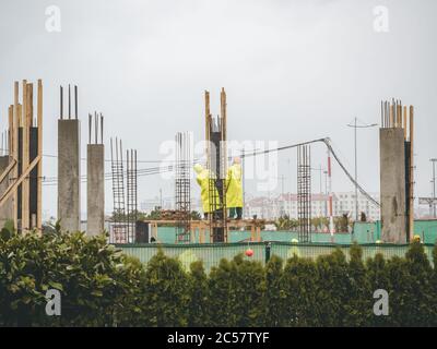I lavoratori in impermeabile giallo si levano in piedi e guardano in su fra le travi dell'edificio circondate da piante verdi contro un cielo nuvoloso. Foto Stock