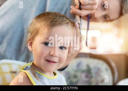 Primo piano giovane adulto caucasica madre che fa pelucchi fot cute adorabile figlio bambino ragazzo a casa a causa quarantena e blocco. Mamma che taglia i capelli del bambino Foto Stock